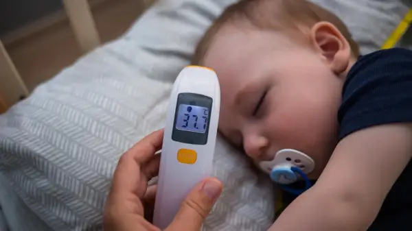 Elektroniczny termometr pokazuje 37.7°C, mierząc temperaturę niemowlaka śpiącego ze smoczkiem.