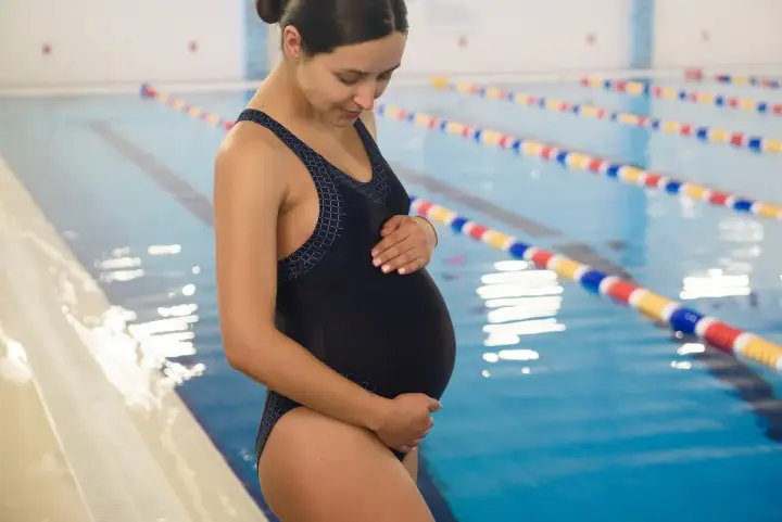 Kobieta w ciąży w czarnym stroju kąpielowym stoi przy basenie, delikatnie dotykając brzucha.